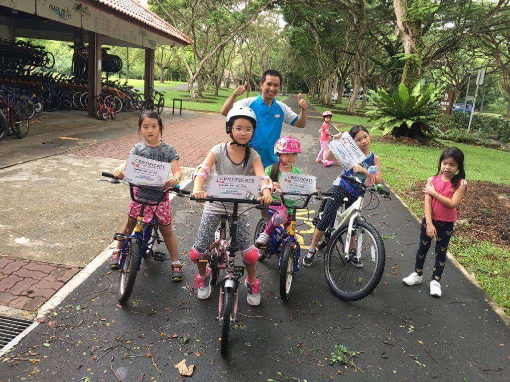 A group of 4 female kids riding a bicycle and holding completion certificate and a male trainer behind them