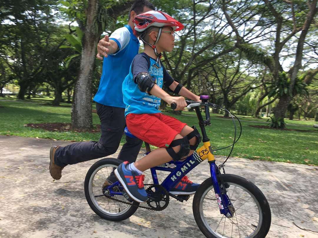 A male kid practicing to ride a bike with a male adult trainer